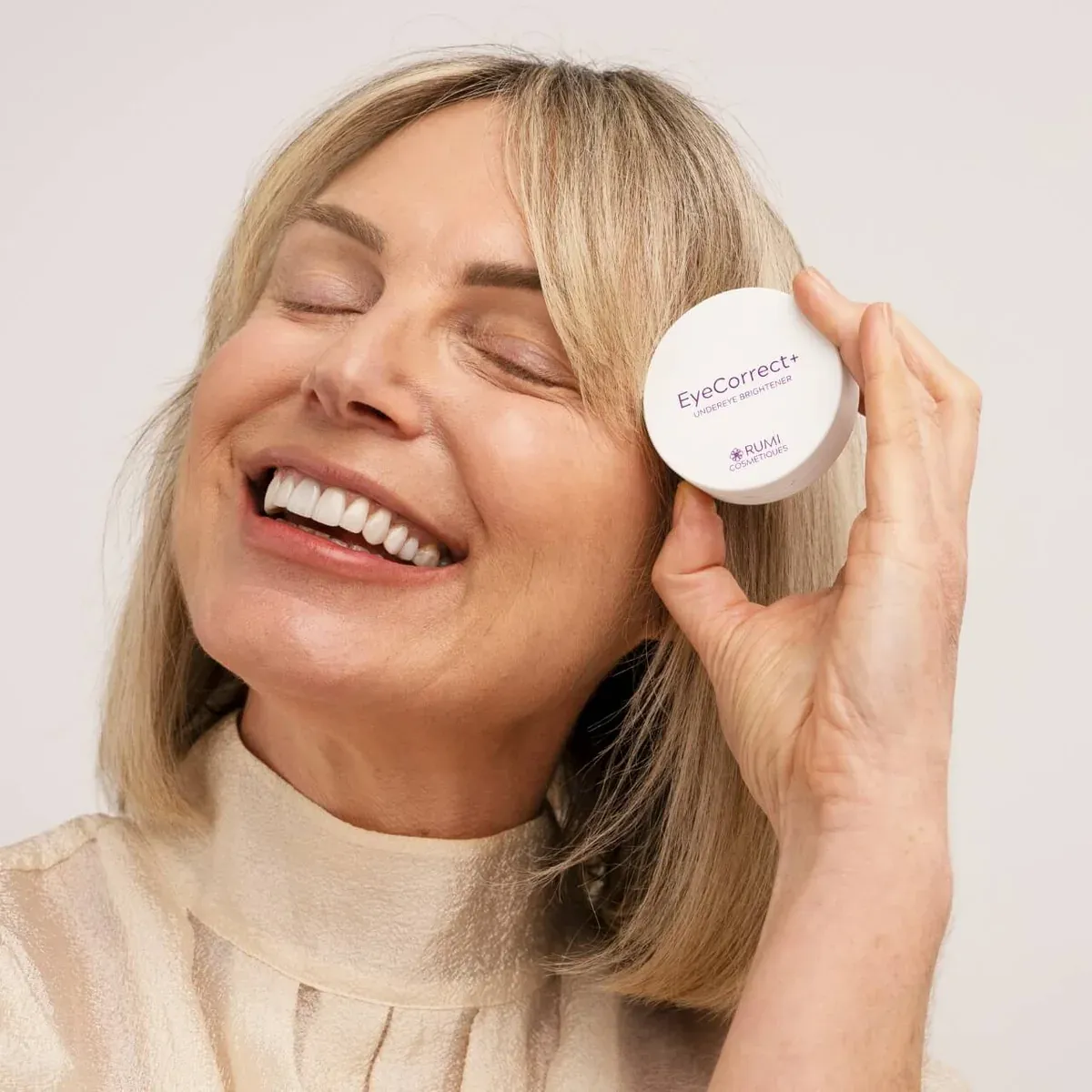 A smiling woman holding a jar of Rumi Cosmetiques EyeCorrect+ undereye brightener beside her face